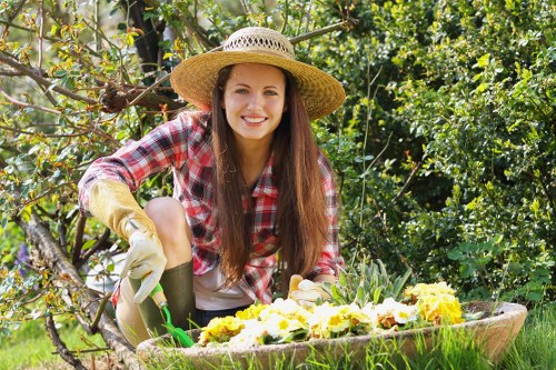A young woman with long brown hair, smiling and wearing a straw hat, plaid shirt, and gardening gloves, is kneeling in a lush garden. She is holding a small gardening trowel and tending to a flower bed filled with yellow and white flowers. Behind her, a dense hedge of greenery and budding trees create a natural backdrop, with sunlight filtering through the leaves. The garden ground features a well-maintained grassy lawn, and nearby, a large wooden planter box contains more flowering plants. In the background, there are tree branches with new growth and a sense of an outdoor, landscaped space typical of gardens in Sutton. The scene conveys active outdoor gardening maintenance, highlighting a healthy garden environment suitable for professional gardening services, with natural earth tones and vibrant plant colours enhancing the visual appeal.