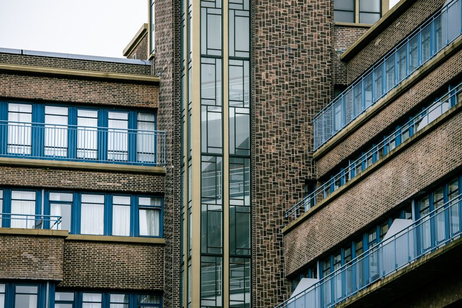 A close-up view of a modern office building with a brick facade, featuring multiple balconies with blue metal railings. The balconies are lined with large, rectangular windows framed in blue, some of