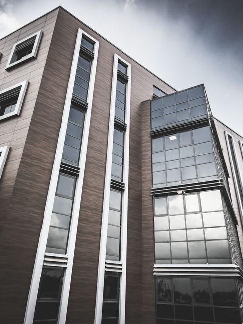 A close-up view of a modern office building with a brick facade, featuring multiple balconies with blue metal railings. The balconies are lined with large, rectangular windows framed in blue, some of