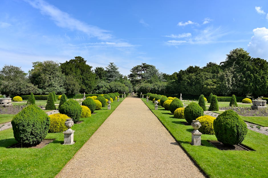 A formal garden scene with a wide, straight gravel pathway running centrally, flanked by symmetrical flower beds with neatly trimmed, rounded bushes and small stone statues or urns placed at intervals