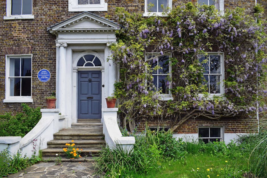 A traditional brick-fronted house with a classic white portico entrance supported by two decorative columns, leading to a dark blue front door. The garden features a stone pathway and steps with yello